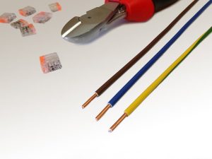 Drei Kupferkabel in Braun, Blau und Gelb-Grün liegen nebeneinander. Oben ist ein Seitenschneider mit rotem Griff zu sehen. Daneben befinden sich mehrere transparente Kabelklemmen. Die Szene vermittelt einen Eindruck von Elektroinstallationsarbeiten.
