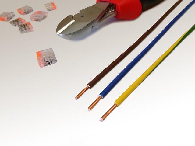 Drei Kupferkabel in Braun, Blau und Gelb-Grün liegen nebeneinander. Oben ist ein Seitenschneider mit rotem Griff zu sehen. Daneben befinden sich mehrere transparente Kabelklemmen. Die Szene vermittelt einen Eindruck von Elektroinstallationsarbeiten.