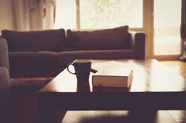 Wohnzimmer mit einem Holztisch im Vordergrund, auf dem eine Tasse und ein Buch liegen; im Hintergrund ein Sofa vor einer großen Fensterfront mit Tageslicht.