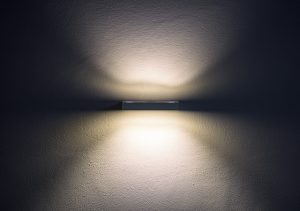 Wandleuchte an einer glatten, dunkelgrauen Wand. Die Lampe strahlt nach oben und unten und erzeugt symmetrische Lichtmuster, die sanft auf der Wand verlaufen. Die Atmosphäre wirkt ruhig und minimalistisch.