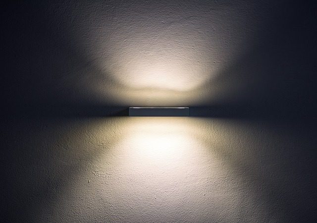 Wandleuchte an einer glatten, dunkelgrauen Wand. Die Lampe strahlt nach oben und unten und erzeugt symmetrische Lichtmuster, die sanft auf der Wand verlaufen. Die Atmosphäre wirkt ruhig und minimalistisch.
