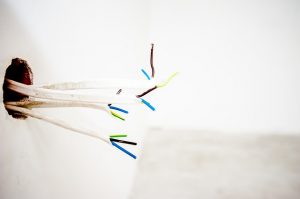 Ein Bündel farbiger Elektrokabel ragt aus einem Loch in der weißen Wand heraus. Die Kabelenden sind unisoliert und zeigen verschiedene Farben wie blau, grün-gelb und braun. Der Hintergrund ist unscharf und schlicht gehalten.