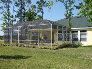 Ein gelbes Haus mit einem anliegenden Wintergarten aus Glas und Metallrahmen. Der Garten ist mit Rasen und einigen Bäumen umgeben. Im Hintergrund sind hohe Bäume sichtbar, die einen klaren, blauen Himmel umrahmen.