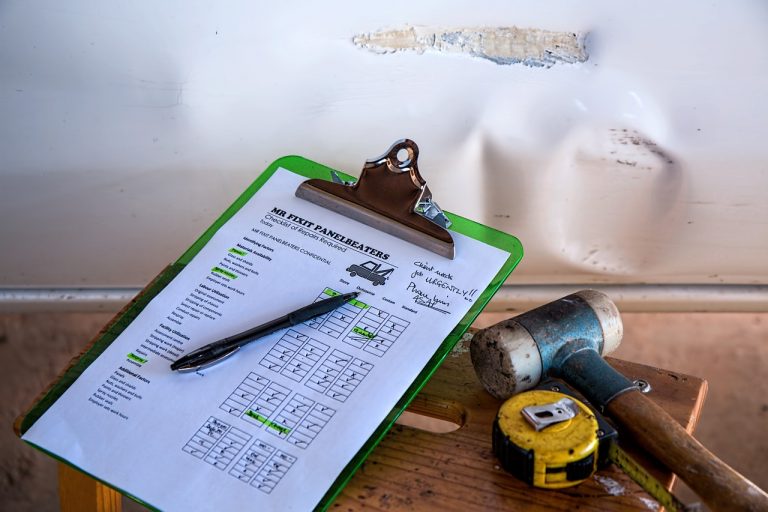 Unfallprotokoll auf einem Klemmbrett mit Kugelschreiber, Maßband und Gummihammer vor einer verbeulten Autotür, Symbol für Schadensaufnahme und Reparaturplanung.
