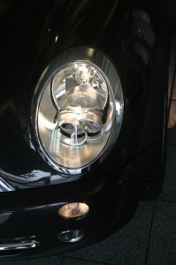 Nahaufnahme eines Autoscheinwerfers an einem dunklen Fahrzeug. Das Licht des Hauptscheinwerfers ist eingeschaltet und strahlt hell. Darunter befindet sich ein kleinerer Nebelscheinwerfer, ebenfalls eingeschaltet. Der Lack des Autos glänzt.