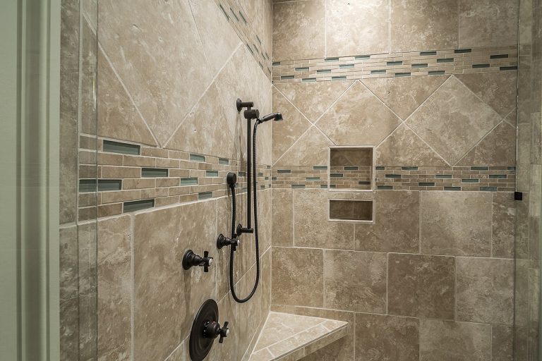 Moderne begehbare Dusche mit beigefarbenen Natursteinfliesen und dekorativen Mosaikbordüren. Die Dusche verfügt über eine schwarze Armatur mit Handbrause, zwei Wandnischen für Duschutensilien und eine gemauerte Sitzbank in der Ecke.