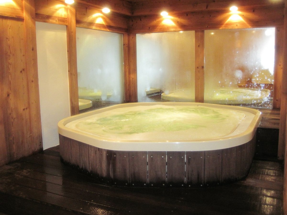 Holzvertäfelter Innenraum mit einem rechteckigen Whirlpool. Warmes Licht von mehreren Deckenstrahlern erzeugt eine gemütliche Atmosphäre. Im Hintergrund sind große, leicht beschlagene Fenster zu sehen.