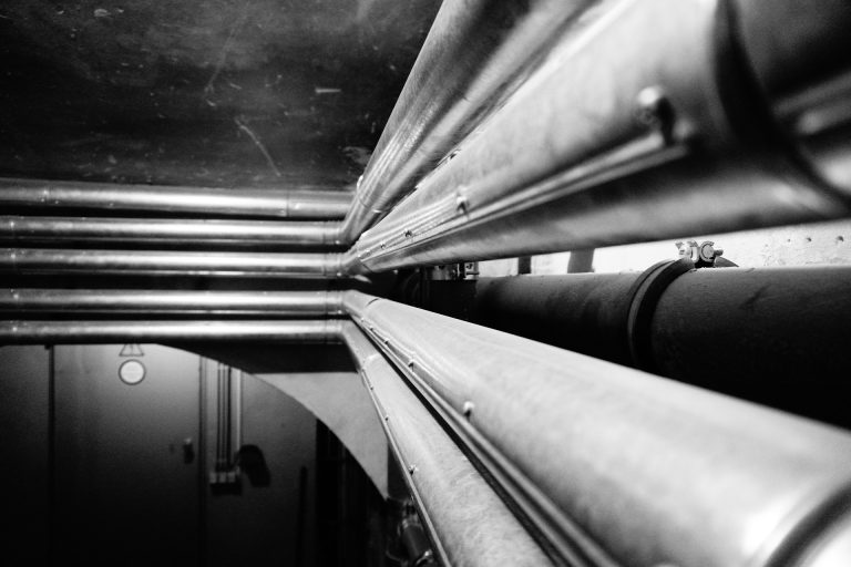 Mehrere parallel verlaufende Heizungsrohre in einem Technikraum oder Kellerbereich mit starker Perspektive in Schwarzweißaufnahme.