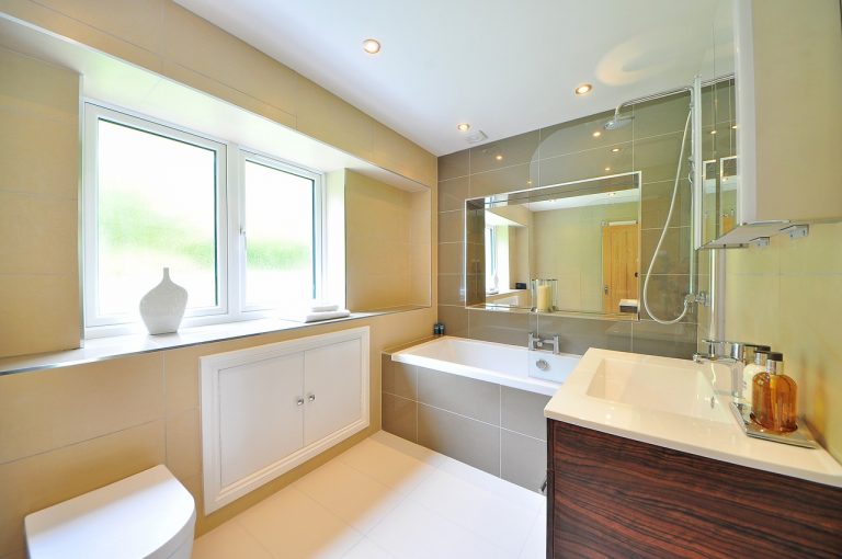 Modernes Badezimmer mit Badewanne, großer Spiegelwand und beiger Wandverkleidung. Die Ausstattung umfasst eine weiße Keramikwanne mit Duscharmatur, ein Waschbecken mit Holzunterschrank und eine breite Fensterbank mit Dekovase.