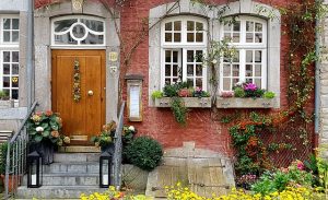 Historische Hausfassade mit dekorativem Eingangsbereich, verziert mit Blumen und traditionellen weißen Sprossenfenstern mit Rundbogen. Die Fenster sind in Naturstein eingefasst und passen stilistisch zur rustikalen roten Ziegelwand.