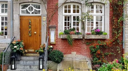 Historische Hausfassade mit dekorativem Eingangsbereich, verziert mit Blumen und traditionellen weißen Sprossenfenstern mit Rundbogen. Die Fenster sind in Naturstein eingefasst und passen stilistisch zur rustikalen roten Ziegelwand.