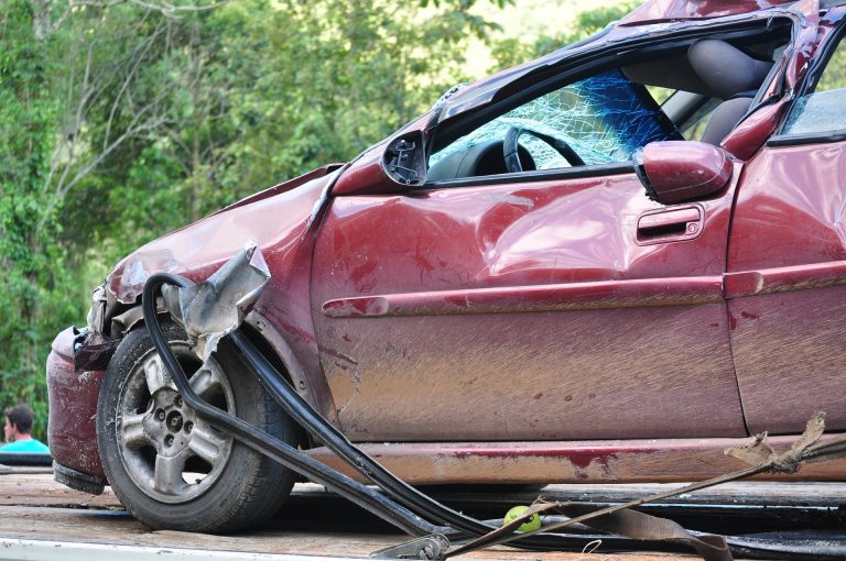 Stark beschädigtes rotes Auto mit eingedrückter Karosserie und zersplitterter Windschutzscheibe nach einem Unfall, auf einem Abschleppfahrzeug vor Waldkulisse.