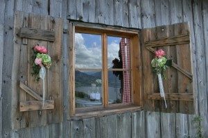 Holzfenster mit offenen Läden, dekoriert mit Blumengestecken. Im Glas spiegelt sich eine Berglandschaft mit blauem Himmel und Wolken. Innen sind rot-weiße Vorhänge sichtbar. Die Szene strahlt ländlichen Charme und Ruhe aus.