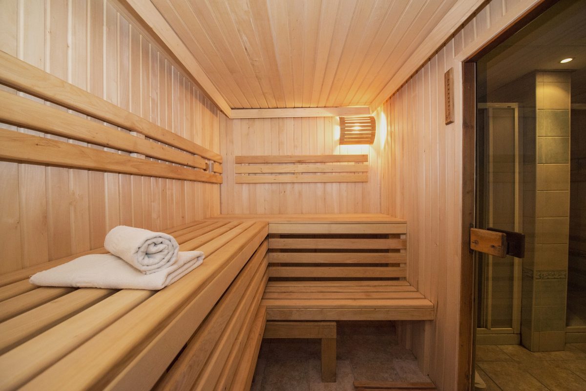 Holzsauna mit Holzbänken und weichen, gefalteten Handtüchern auf der rechten Bank. Die Wände und Decke sind aus hellem Holz. Ein weiches Licht schafft eine warme und entspannende Atmosphäre.