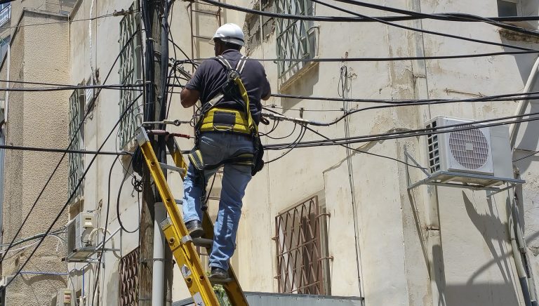 Ein Elektriker steht auf einer Leiter an einem Strommast und arbeitet an einem Geflecht aus Strom- und Datenleitungen. Er trägt Schutzkleidung mit Helm und Klettergurt. Die Wand im Hintergrund ist alt und mit mehreren Klimaanlagen ausgestattet. Das Bild thematisiert den dichten Kabelverlauf und mögliche Belastung durch Elektrosmog.