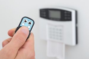 Nahaufnahme einer Hand, die eine Fernbedienung mit zwei Knöpfen hält. Im Hintergrund ist ein an der Wand montiertes Sicherheitssystem mit einem kleinen Bildschirm und Tasten zu sehen. Die Fernbedienung zeigt Symbole eines geschlossenen und eines offenen Schlosses.
