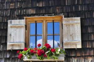 Rustikales Holzfenster mit Fensterläden und Sprossen, geschmückt mit roten Geranien in einem Blumenkasten vor einer dunklen Holzschindelfassade.