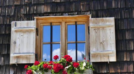 Rustikales Holzfenster mit Fensterläden und Sprossen, geschmückt mit roten Geranien in einem Blumenkasten vor einer dunklen Holzschindelfassade.