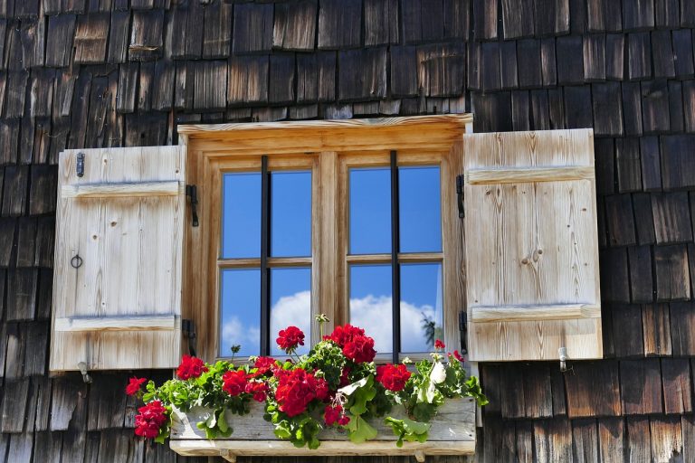 Rustikales Holzfenster mit Fensterläden und Sprossen, geschmückt mit roten Geranien in einem Blumenkasten vor einer dunklen Holzschindelfassade.