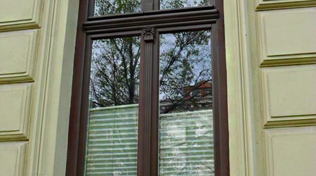 Klassisches Holzfenster mit aufgesetzter Zierleiste in einem Altbau, bestehend aus zwei senkrechten Flügeln und historischer Sprossenteilung, ergänzt durch innenliegende Jalousien.