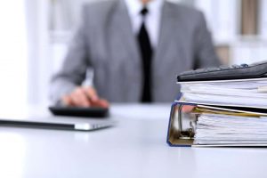 Ein Mann in einem grauen Anzug sitzt an einem Schreibtisch. Im Vordergrund liegen ein Stapel Aktenordner und ein Taschenrechner. Der Fokus liegt auf den Dokumenten, während die Person im Hintergrund unscharf dargestellt ist.
