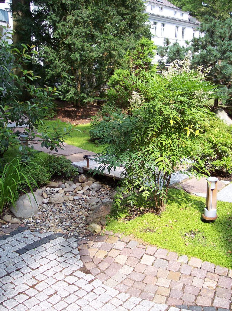 Gepflasterter Gartenweg mit kleinem Bachlauf, umgeben von dichten Sträuchern und Bäumen in einer gepflegten Gartenanlage.
