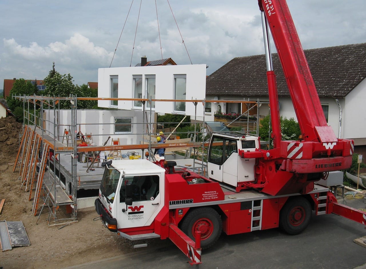 Montage eines Fertighauses auf einer Baustelle mit rotem Mobilkran, der ein vorgefertigtes Wandelement einsetzt, umgeben von Baugerüsten und Bauarbeitern.