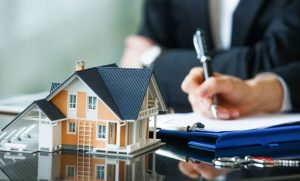 Ein Modellhaus steht auf einem Tisch, während eine Person im Hintergrund Papiere unterzeichnet. Neben dem Modell liegen Schlüssel und ein Klemmbrett. Die Szene vermittelt den Eindruck eines Immobiliengeschäfts oder eines Hauskaufs.