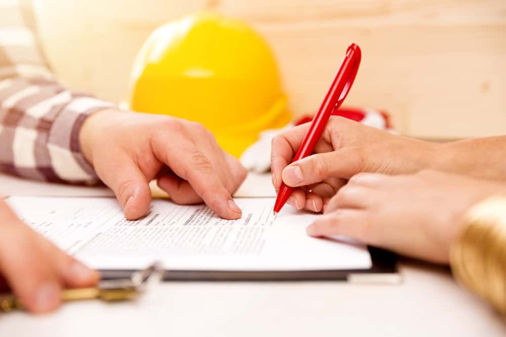 Zwei Personen sitzen an einem Tisch und arbeiten an einem Dokument. Eine Person zeigt mit einem Finger auf den Text, während die andere mit einem roten Stift schreibt. Im Hintergrund ist ein gelber Bauhelm zu sehen.