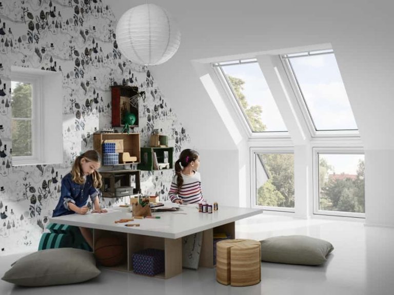 Zwei Mädchen malen an einem weißen Tisch in einem hellen Raum mit großen Dachfenstern. Die Wand ist mit schwarz-weißen Mustern verziert. Der Raum hat Kissen und Regale mit bunten Gegenständen und Spielsachen.