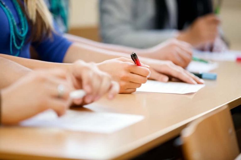 Mehrere Personen sitzen an einem Tisch und schreiben auf Papier. Sie verwenden verschiedene Stifte in einem Klassenzimmer oder einer Schulungsumgebung. Der Fokus liegt auf den Händen der Personen und dem Schreibprozess.