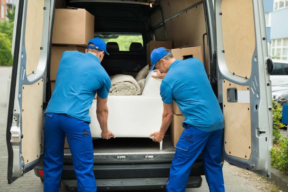 Zwei Männer in blauen Uniformen tragen ein Möbelstück in einen offenen Lieferwagen. Im Hintergrund sind Kartons und ein zusammengerollter Teppich sichtbar. Die Szene spielt sich auf einem Parkplatz vor einem Gebäude ab.