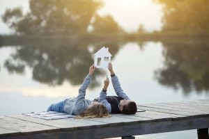 Zwei Personen liegen auf einem Steg am See und halten gemeinsam einen weißen Umriss eines Hauses in die Luft. Der See reflektiert die umgebende Natur und die Szene ist in warmes, sanftes Licht getaucht.