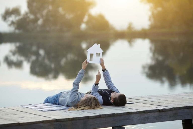 Zwei Personen liegen auf einem Steg am See und halten gemeinsam einen weißen Umriss eines Hauses in die Luft. Der See reflektiert die umgebende Natur und die Szene ist in warmes, sanftes Licht getaucht.