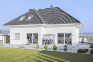 Modernes weißes Einfamilienhaus mit Walmdach, großen Fensterfronten und gepflegtem Garten bei klarem Himmel.