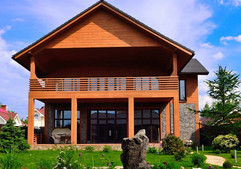 Modernes zweistöckiges Holzhaus mit Naturstein-Fundament, großer Glasfassade und gepflegtem Garten unter blauem Himmel.
