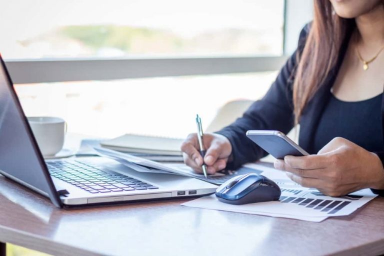 Frau arbeitet am Laptop, hält ein Smartphone in der Hand und macht sich Notizen.