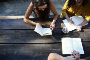 Drei Personen sitzen an einem Holztisch. Zwei von ihnen lesen Bücher, während die dritte Person in ein Notizbuch schreibt. Ein Getränk im Plastikbecher steht auf dem Tisch. Alle scheinen konzentriert zu arbeiten.