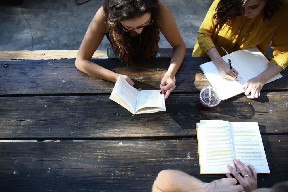 Drei Personen sitzen an einem Holztisch. Zwei von ihnen lesen Bücher, während die dritte Person in ein Notizbuch schreibt. Ein Getränk im Plastikbecher steht auf dem Tisch. Alle scheinen konzentriert zu arbeiten.