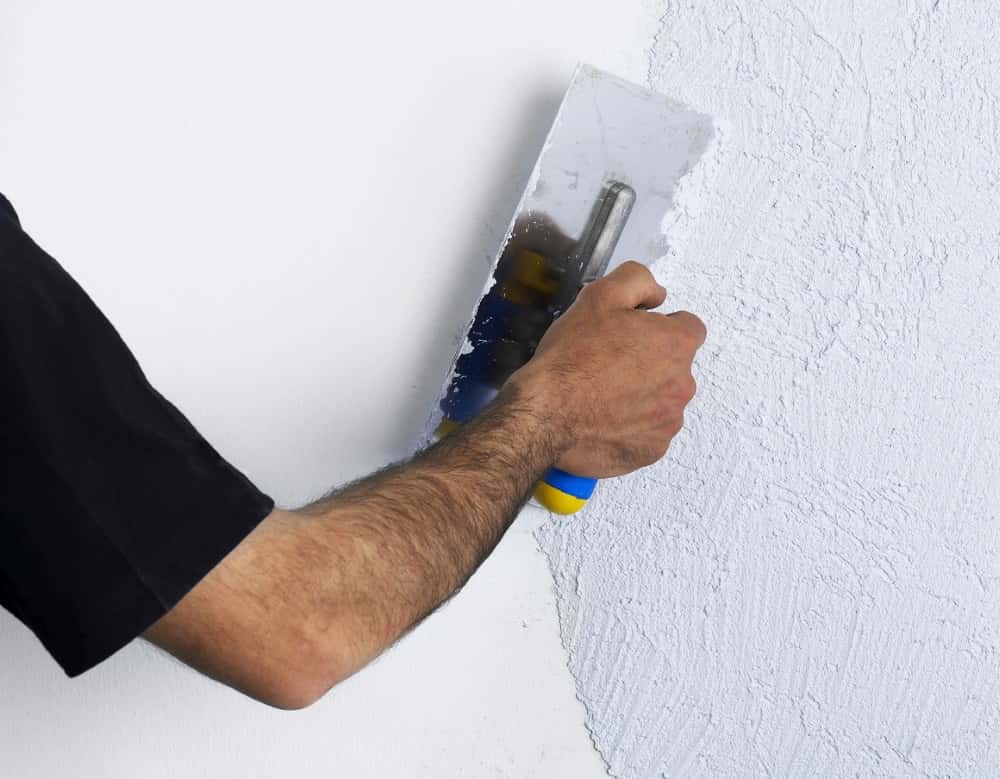 Ein Mensch verputzt eine weiße Wand mit einer Kelle. Die Wand hat bereits eine strukturierte Oberfläche. Die Person trägt ein schwarzes T-Shirt.