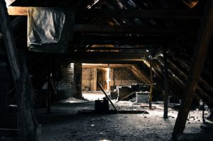 Dunkler, verlassener Dachboden mit Holzbalken und teilweise eingestürztem Dach. Schwaches Licht fällt durch Ritzen, beleuchtet Staub und Trümmer. Im Hintergrund ist eine zugesperrte Holztür zu sehen, die zu einem weiteren Raum führt.