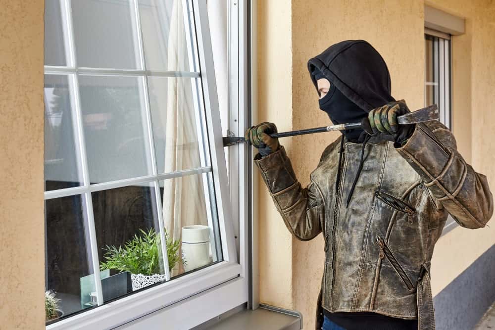 Maskierter Einbrecher versucht mit einem Brecheisen, ein Fenster eines Wohnhauses aufzubrechen – Darstellung eines Einbruchs oder Sicherheitsrisikos im Eigenheim.