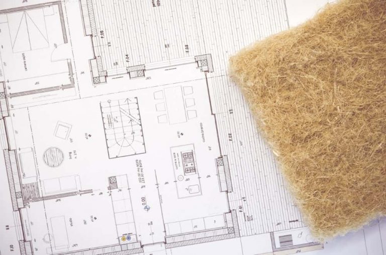 Ein Architekturplan liegt auf einem Tisch, daneben befindet sich ein Stück Dämmmaterial in brauner Farbe. Der Grundriss zeigt verschiedene Räume und Details zu einem Wohnbereich mit Möbelplatzierungen.