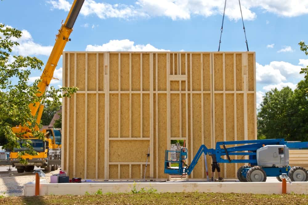 Kran hebt eine große Holzwand auf einer Baustelle. Ein Arbeiter steht auf einer blauen Hebebühne und arbeitet an der Wand, umgeben von grünen Bäumen. Baumaterialien liegen am Boden vor der Struktur. Blauer Himmel mit wenigen Wolken.