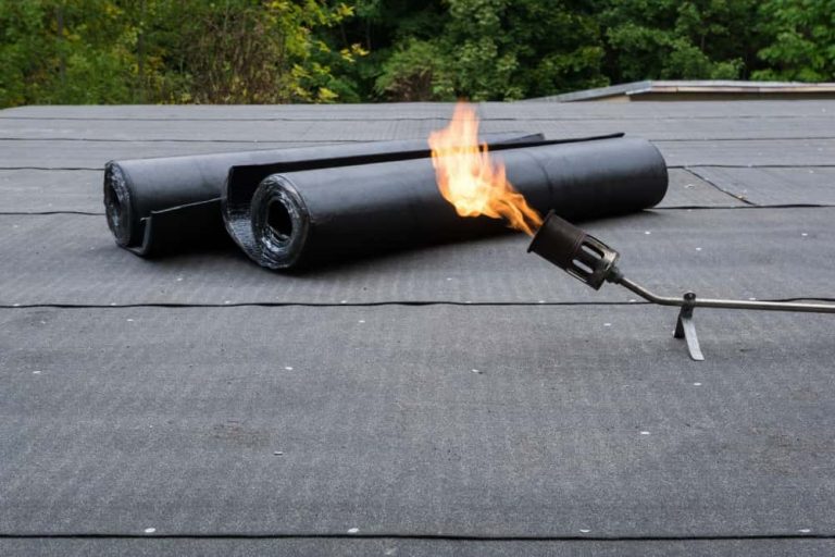 Flachdach mit zwei Rollen Dachpappe und einem Brenner, dessen Flamme in die Luft ragt. Der Hintergrund zeigt grüne Bäume, die das Bild umrahmen. Die Szene deutet auf Arbeiten zur Abdichtung oder Reparatur der Dachfläche hin.