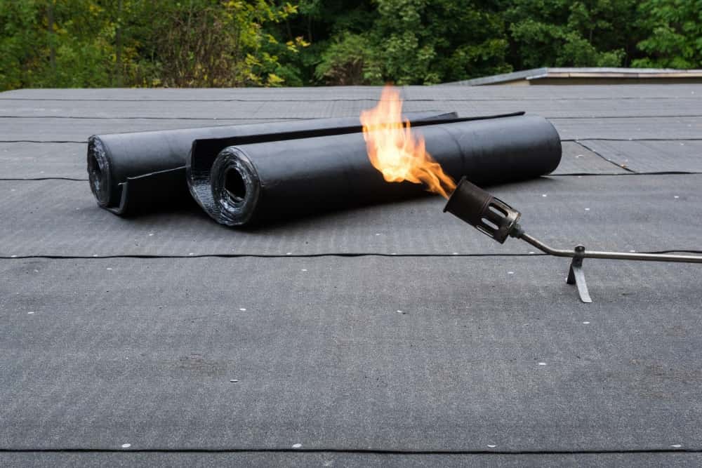 Flachdach mit zwei Rollen Dachpappe und einem Brenner, dessen Flamme in die Luft ragt. Der Hintergrund zeigt grüne Bäume, die das Bild umrahmen. Die Szene deutet auf Arbeiten zur Abdichtung oder Reparatur der Dachfläche hin.