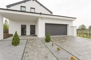 Modernes Einfamilienhaus mit Flachdach-Anbau, grauer Doppelgarage und minimalistischer Vorgartengestaltung mit Kiesflächen und kleinen Ziersträuchern.