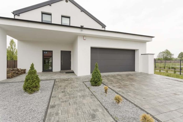 Modernes Einfamilienhaus mit Flachdach-Anbau, grauer Doppelgarage und minimalistischer Vorgartengestaltung mit Kiesflächen und kleinen Ziersträuchern.