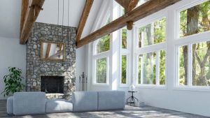 Modern eingerichtetes Wohnzimmer mit großen weißen Sprossenfenstern, freiliegenden Holzbalken, Naturstein-Kamin und hellgrauem Sofa in offenem, lichtdurchflutetem Raum mit Waldblick.