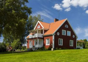 Rotes Schwedenhaus mit weißem Balkon, Satteldach und Holzfassade, umgeben von Rasen, Bäumen und ländlicher Natur unter blauem Himmel.